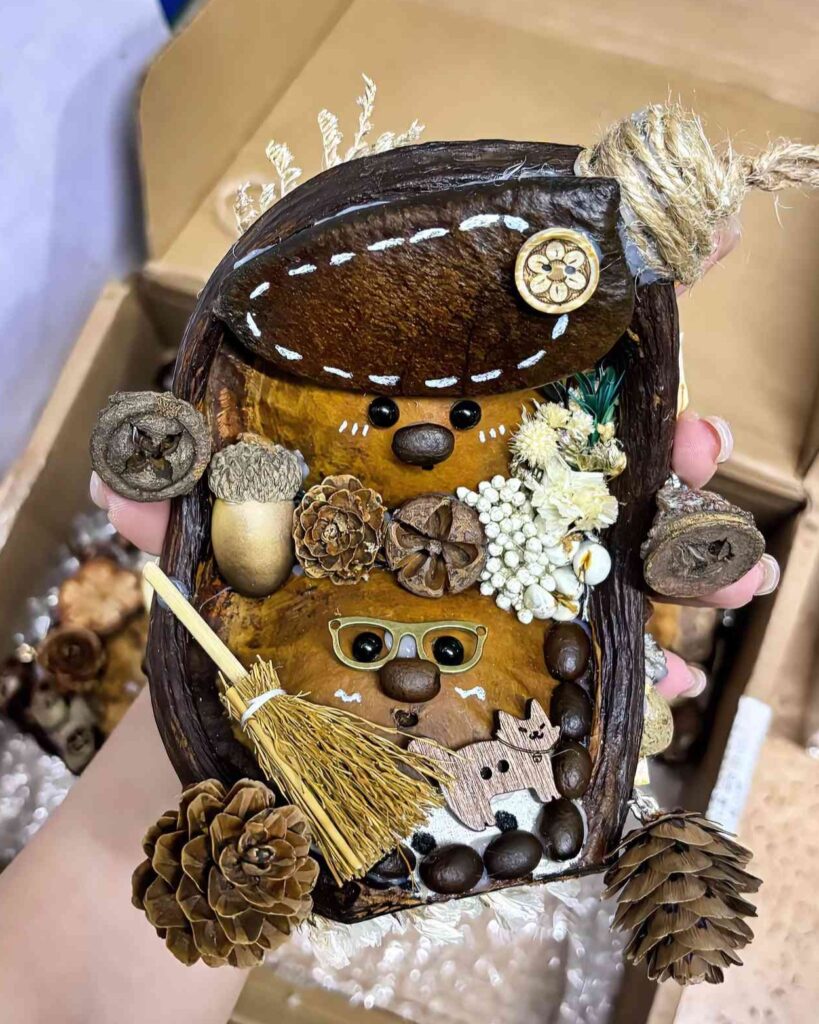 A cute handmade ornament featuring two tiny characters crafted from Entada seed pods, decorated with natural elements like pinecones, dried flowers, coffee beans, and a straw broom. Designed with playful details and earthy textures, this hanging charm is perfect as a car accessory or a thoughtful gift for nature lovers.