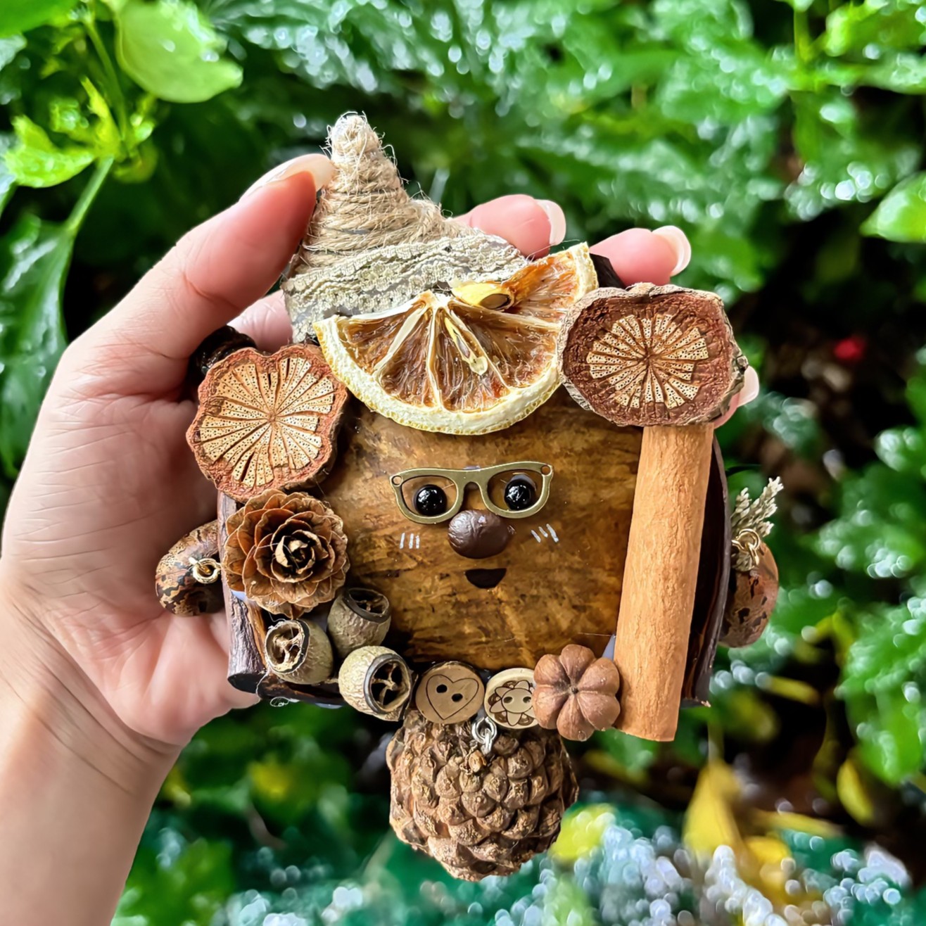 A cute handmade ornament made from natural Entada seed pod, decorated with dried citrus, cinnamon stick, pinecones, and seed shells. This whimsical pendant features a smiling face with glasses and is perfect as a creative gift or a cozy car hanging charm.